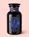 A black glass bottle featuring a geometric blue label, inspired by Sapphire: Apple-Plum-Spice White Tea for Self-Expression, and adorned with an ornate neck design rests against a pale pink background, evoking a sense of spiritual wellness.