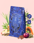 A Sapphire: Apple-Plum-Spice White Tea for Self-Expression package, featuring a gemstone-inspired design, is displayed against a pale pink background and surrounded by fresh ingredients—lemongrass, butterfly pea flowers, papaya, cherry, apple slices, cinnamon sticks, and green herbs. This grounding white tea blend invites tranquility and encourages natural wellness.