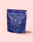 A blue resealable pouch with a geometric gemstone design and "Gemstone Wellness Tea, Sapphire" label sits on a pale pink background. Sapphire: Apple-Plum-Spice White Tea for Self-Expression highlights the elegance of organic white tea.