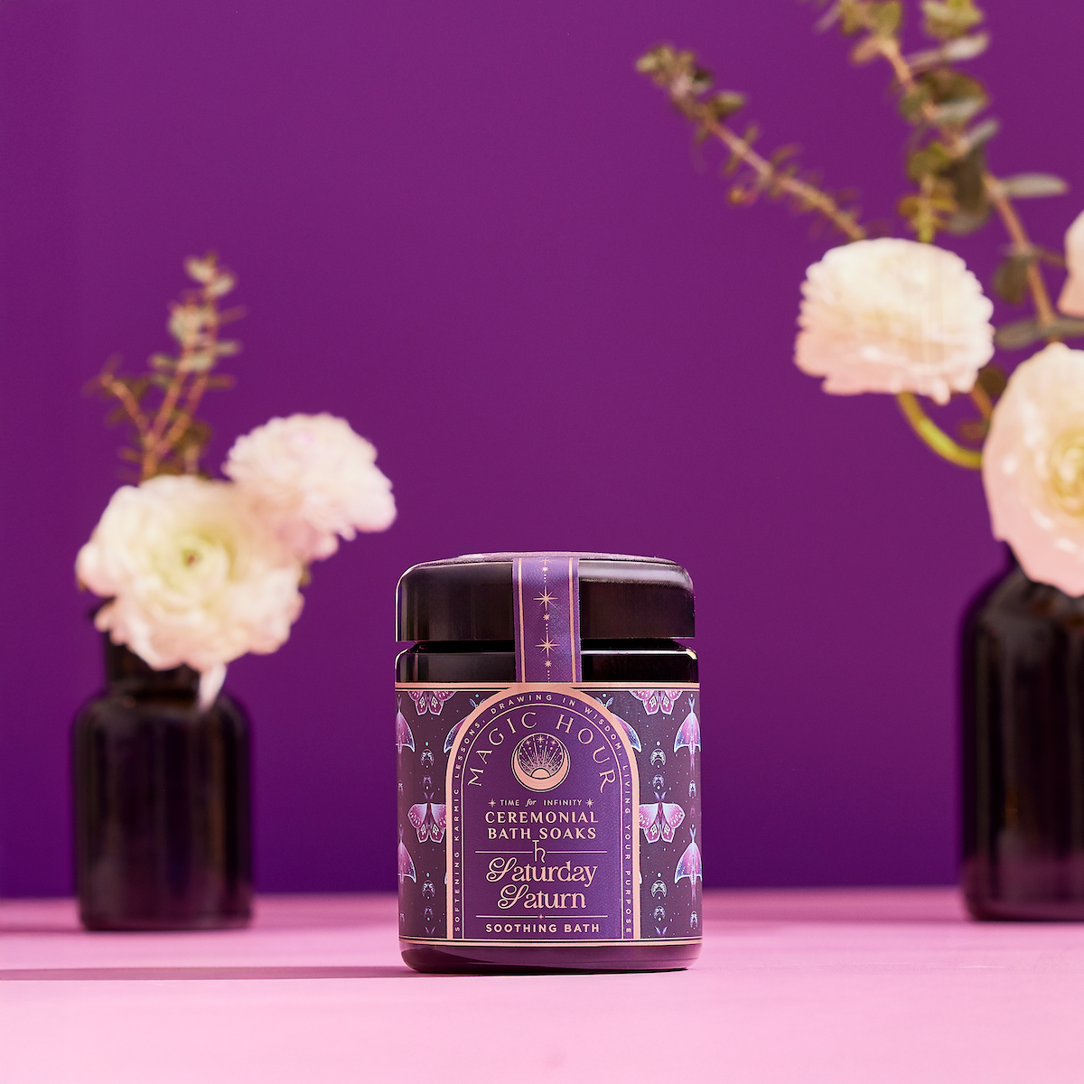 A jar of Ceremonial Bath Salt Soaks in a violet glass jar is centered on a pink surface with a purple backdrop, ideal for self-care rituals that inspire a celestial connection, flanked by blurred vases of white flowers.