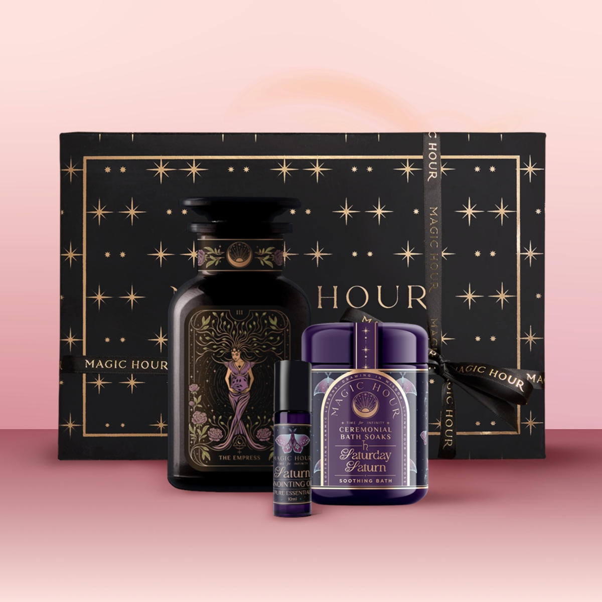 The Saturn Bath & Beauty Box features a sacred beauty box, a large black jar with mystical design, a small roll-on, and a purple container, all arranged on a pink surface to inspire grounding energy.
