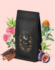 A black coffee bag with a floral label reading "Scorpio Tea for Sensual Brilliance" is surrounded by roses, cinnamon sticks, figs, and other flowers on a light pink background, evoking the delicate allure of rose congou and the enchanting notes of sensual tea.