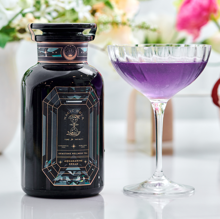 A decorative dark bottle labeled "Aquamarine Dream - Soothing Herbal Ayurvedic Adrenal Tonic" stands next to a glass of vibrant purple liquid, with blurred flowers in the background on a white surface.