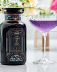 A decorative dark bottle labeled "Aquamarine Dream - Soothing Herbal Ayurvedic Adrenal Tonic" stands next to a glass of vibrant purple liquid, with blurred flowers in the background on a white surface.