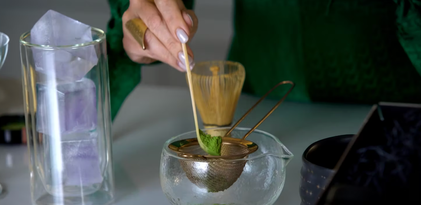 A person in a green outfit is scooping matcha powder with a bamboo spoon and placing it into a metal strainer over a glass teapot. Nearby are a bamboo whisk, a glass of ice, and other tea-making utensils.