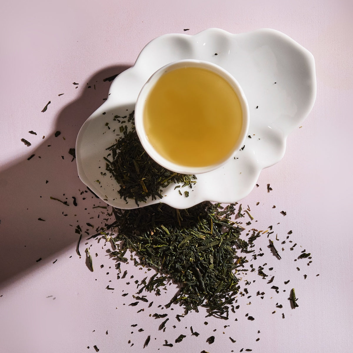 A white ceramic teacup filled with light Sencha Kyoto Green Tea rests on a uniquely shaped white saucer. Loose green tea leaves, rich in antioxidants and known to boost metabolism, are scattered around the saucer and teacup on a soft pink background. The image is well-lit, highlighting the texture of the leaves and the tea&#39;s color.