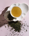 A white ceramic teacup filled with light Sencha Kyoto Green Tea rests on a uniquely shaped white saucer. Loose green tea leaves, rich in antioxidants and known to boost metabolism, are scattered around the saucer and teacup on a soft pink background. The image is well-lit, highlighting the texture of the leaves and the tea's color.