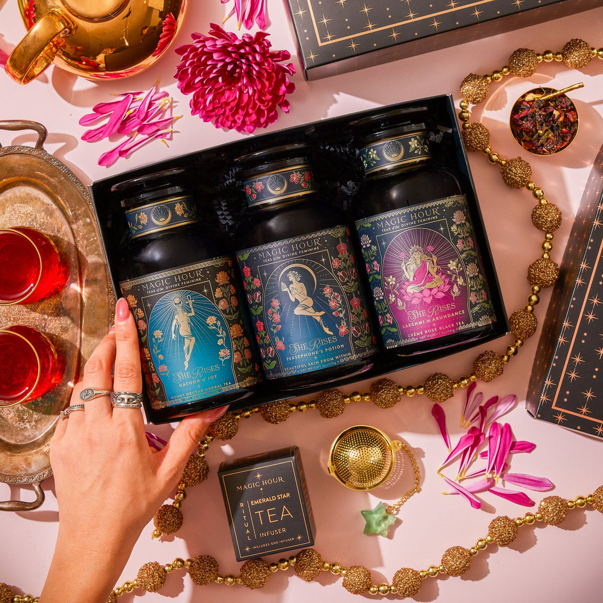 A She Rises Goddess Golden Hour Apothecary Gift Box containing three apothecary tea jars, surrounded by a gold teapot, two glasses of red tea, scattered flower petals, and a gold tea infuser.