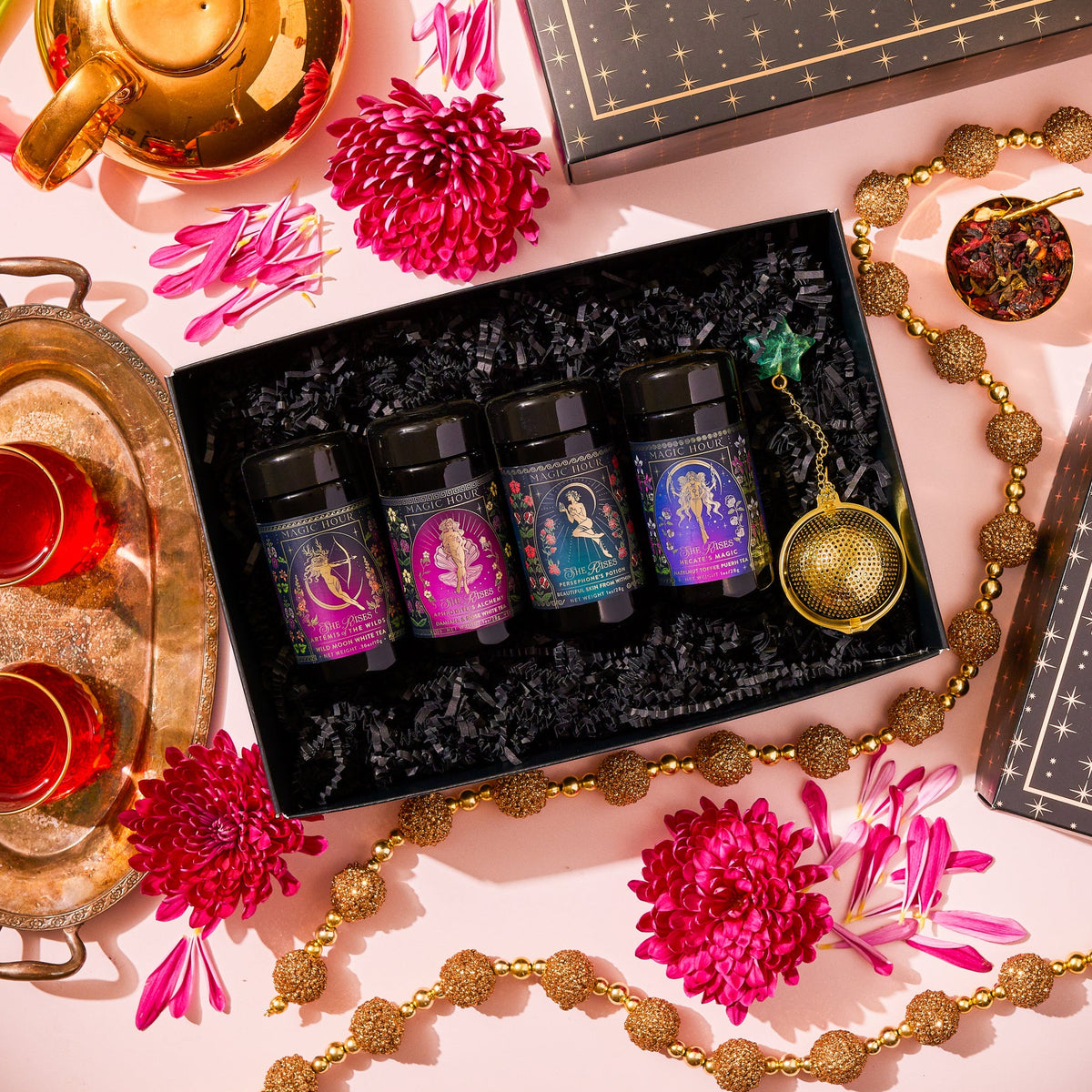 A She Rises Goddess Traveler Gift Box containing four traveler tea jars and an Emerald Star Strainer, surrounded by gold beads, pink flower petals, two glasses of tea, and a tea kettle.