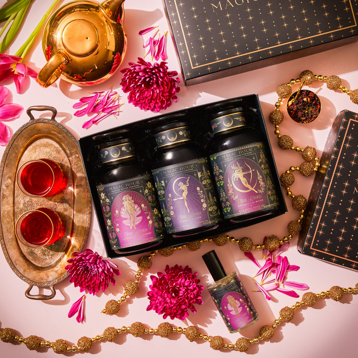 A She Rises Goddess Moonlit Bloom Apothecary Gift Box containing three apothecary jars of moonlight white tea, a spray bottle, and a gold teapot, surrounded by pink flowers, a tray with two cups of tea, and beaded garlands.