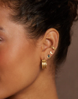 A close-up of a personalized ear stack adorned with multiple gold earrings and the Magic Hour Shine On Beaded Ear Cuff, including a small hoop with a sun charm, a crescent moon stud, a small bi-color stud, and a larger textured hoop earring at the lobe. The background is plain brown.