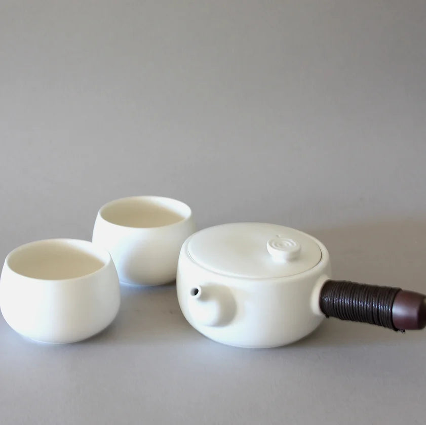 A minimalist Side Handle Tea Set by Eastern Elm, featuring a white ceramic teapot with a cylindrical brown handle and two round tea cups placed beside it on a plain, light grey background. The set combines traditional craftsmanship with modern, sleek design.