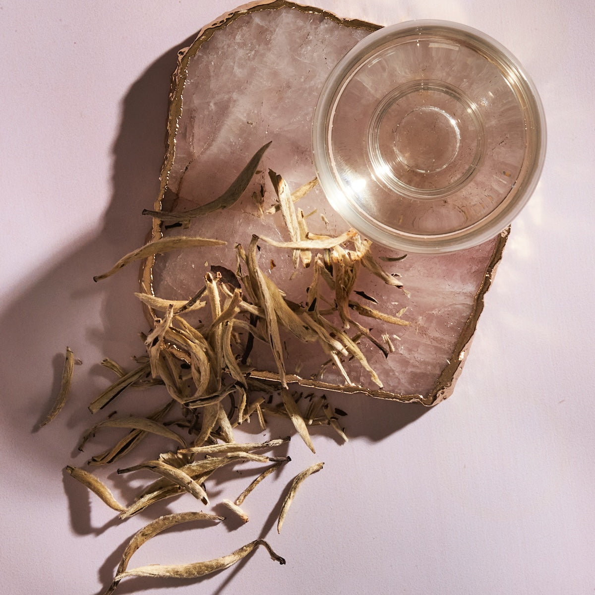 A glass cup of clear liquid sits on a polished pink stone slab with metallic gold edges. Dried Club Magic Hour Silver Moon White Tea leaves are scattered on the surface of the slab and the surrounding area. Shadows of the cup and tea leaves fall on the light pink background.
