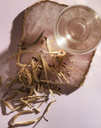 A glass cup of clear liquid sits on a polished pink stone slab with metallic gold edges. Dried Club Magic Hour Silver Moon White Tea leaves are scattered on the surface of the slab and the surrounding area. Shadows of the cup and tea leaves fall on the light pink background.