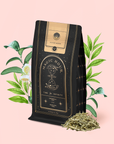 A black and gold Magic Hour tea bag labeled "Silver Moon White Tea: Yin Zhen Silver Needle" stands on a pink background, with loose Fujian white tea leaves and illustrated green tea plants and flowers beside it.