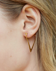 Close-up of a woman's ear wearing Magic Hour's Simple Strength Earrings, a delicate, 18k gold V-shaped earring. The modern silhouette dangles slightly below the ear lobe, while her light brown hair partially covers the ear. The background is blurred, keeping the focus on the ear and earring.