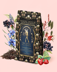 A decorative tea bag labeled "Skadi The Sovereign: Winter Berry Puerh" is surrounded by illustrated berries, flowers, leaves, and loose tea leaves on a pink background—evoking the enchanting spirit of the Norse Winter Queen and her winter resilience with every sip of this berry-infused puerh tea.