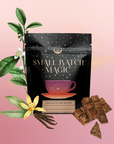 A black pouch labeled "Small Batch Magic Valentine’s 2026" with a teacup illustration rests among vanilla flowers, cocoa leaves, and chocolate squares, evoking notes of chocolate cake tea and Madagascar vanilla rooibos on a pink gradient background.