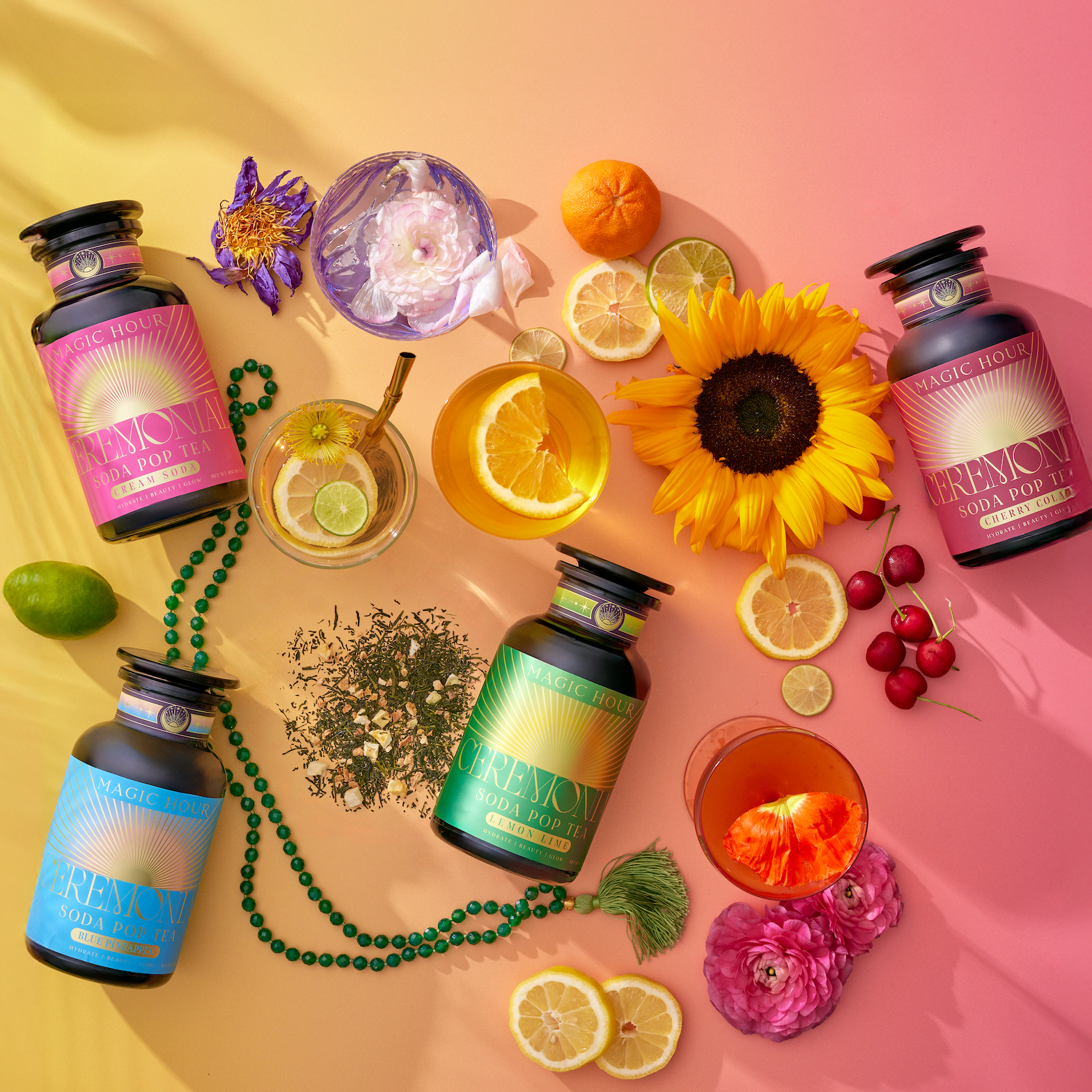 Bottles of the Ceremonial Soda Tea Bundle, a caffeine-free treat, are displayed with a sunflower, citrus slices, flowers, herbs, cherries, beads, and drinks on a pink and yellow gradient background.
