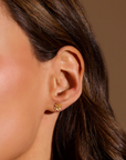 Close-up of a person's right ear adorned with a small, Magic Hour Soul Renewal Gold Lotus Earring featuring white topaz. The person has long, brown hair, and the background is a warm neutral tone. The delicate earring symbolizes new beginnings and complements the person's skin tone beautifully.