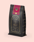 The Soulmate Chocolate-Raspberry-Rose Black Tea refill pouch features a celestial black and gold design labeled "Magic Hour, Time for Infinity." It includes an hourglass and stars, evoking wellness rituals. A red and gold top contrasts elegantly with the soft pink background.