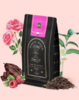 A black refill pouch of Soulmate: Chocolate-Raspberry-Rose Black Tea with a pink label is surrounded by illustrated roses, cacao pods, raspberries, and tea leaves on a pale pink background.