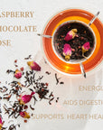 A glass cup of tea with loose leaf tea leaves, rose petals, and a tea infuser sits on a white surface. The tea is a rosy-brown color. Text around the image reads: "SOULMATE: CHOCOLATE-RASPBERRY-ROSE BLACK TEA FOR FINDING & CELEBRATING LOVE," "ENERGIZES," "AIDS DIGESTION," and "SUPPORTS HEART HEALTH." Experience the magic hour with organic tea bliss from Magic Hour.