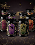 Four glass jars from the Spellbound Sips Bundle arranged in a semicircle, featuring ornate black labels with gold illustrations and text, set against a dark background with gold star and butterfly accents.