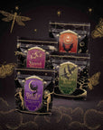 Four black pouches of Spellbound Sips Bundle tea, decorated with gold celestial and nature illustrations and labeled “Haunted Hibiscus,” “Candy Corn,” “Kettle Corn Kiss,” and “Black Cat Brownie.”.