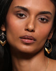 A person with long, dark hair is wearing the exquisite Spring Lotus Earrings, made of 18kt gold plating and shaped like leaves. Adorned with blue topaz, these earrings add a touch of elegance. With a neutral expression, the person looks directly at the camera against a plain, light-colored background.