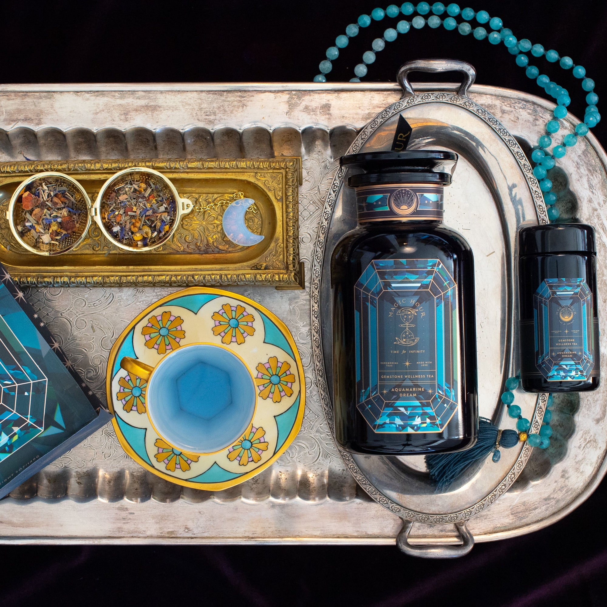A silver tray holds an Aquamarine Dream tea tin, a blue and gold teacup, a large and small dark blue canister, a turquoise beaded tasbih, and a gold tray with two cups of dried tea featuring a moon design.