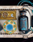 A silver tray holds an Aquamarine Dream tea tin, a blue and gold teacup, a large and small dark blue canister, a turquoise beaded tasbih, and a gold tray with two cups of dried tea featuring a moon design.
