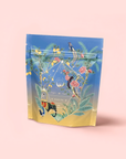 A stand-up pouch of Sri Lankan Sunrise breakfast tea features a vibrant illustrated design with tropical birds, green leaves, and pink flowers on a gradient blue and yellow background, set upon a soft pink surface.