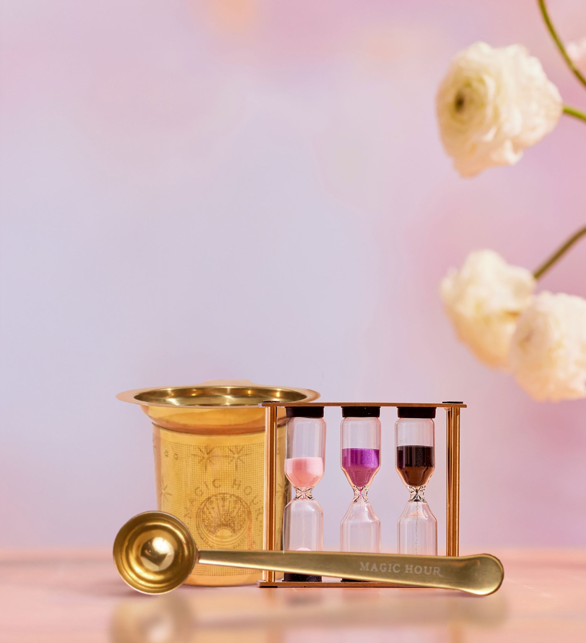 The Loose Leaf Tea Starter Set includes a gold tea strainer, a "MAGIC HOUR" gold spoon, and three colored-sand tea timer hourglasses on a pink surface with white flowers and a pastel background.