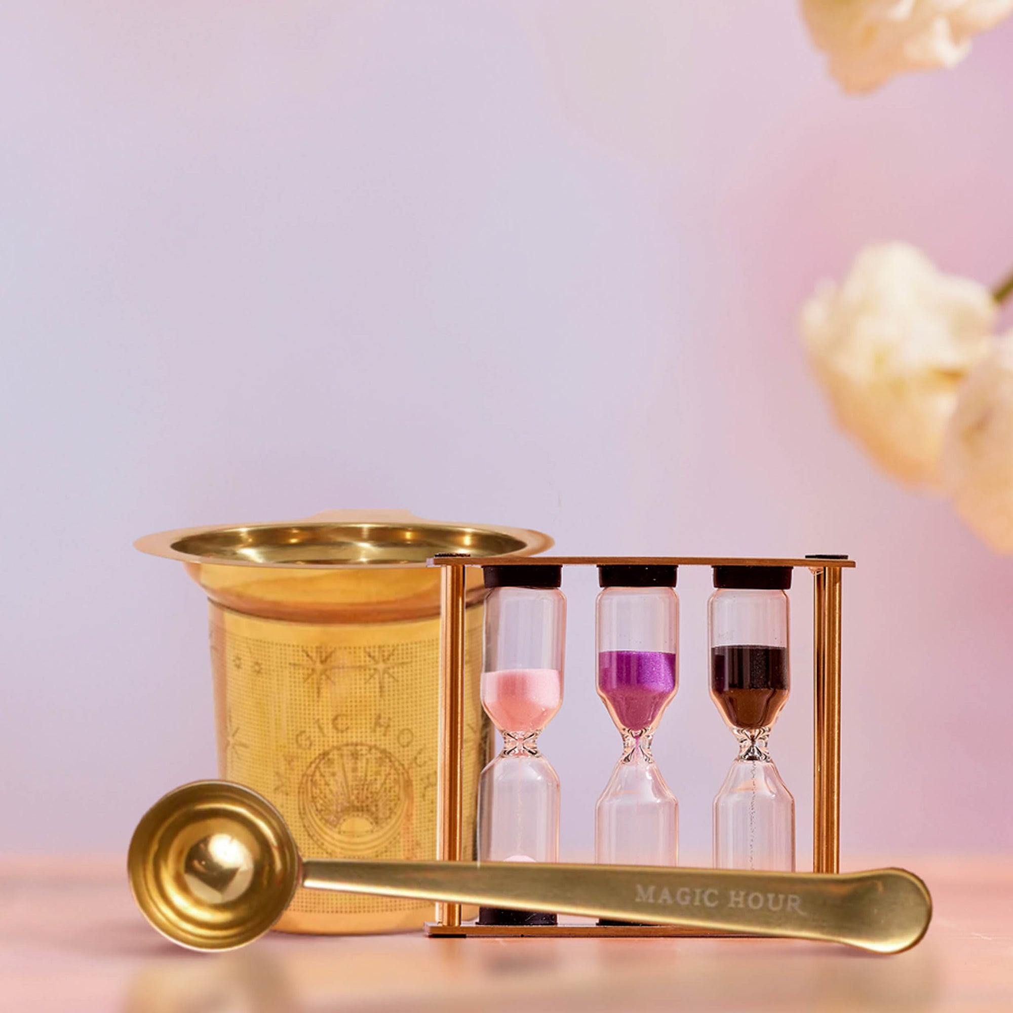 Loose Leaf Tea Starter Set: Includes a gold tea strainer, matching spoon, and a three-hourglass tea timer with pink, purple, and brown sands—all beautifully arranged on a light surface with blurred flowers in the background. This elegant set is perfect for brewing your favorite loose-leaf tea.
