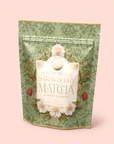 A green floral pouch labeled “Flower Powder: Strawberry Matcha”—adorned with strawberries and white flowers—stands on a light pink background, inviting you to experience the wellness benefits of this premium ceremonial matcha blend.