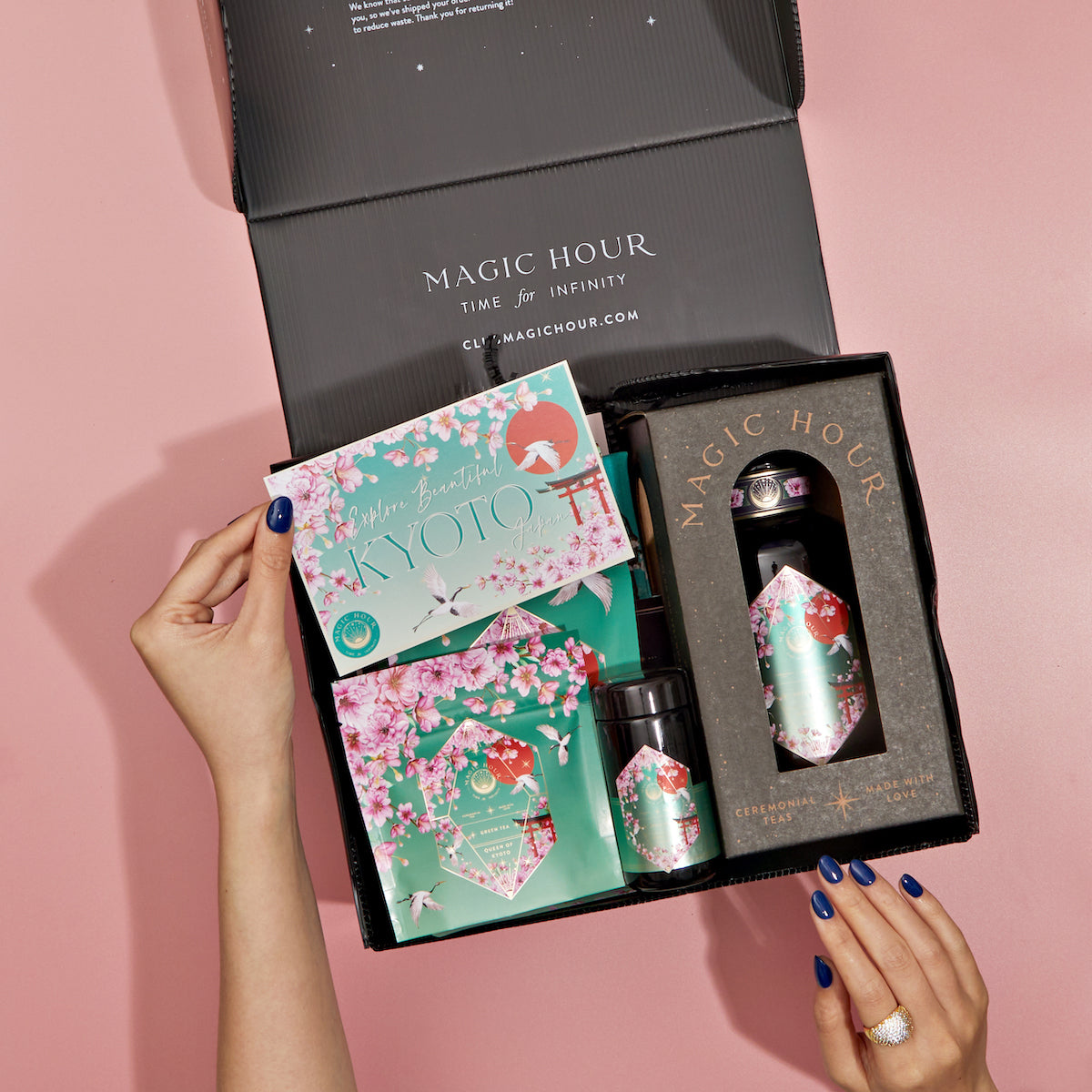 A person opens a Monthly Magic 3-Month Tea Subscription Box, unveiling a selection of Kyoto-themed tea products. The packaging is adorned with cherry blossoms and a teal color scheme. With nails painted blue, they savor the herbal teas, while a ring on one hand glimmers in the light.