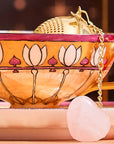 A close-up shot features a decorative teacup and saucer adorned with pink and gold patterns. The Rose Quartz Heart Tea Strainer—a gold tea infuser accented with a heart-shaped rose quartz stone charm—dangles gracefully from the cup’s handle, making it an ideal accessory to elevate any tea ritual with the gentle, loving energy of rose quartz.