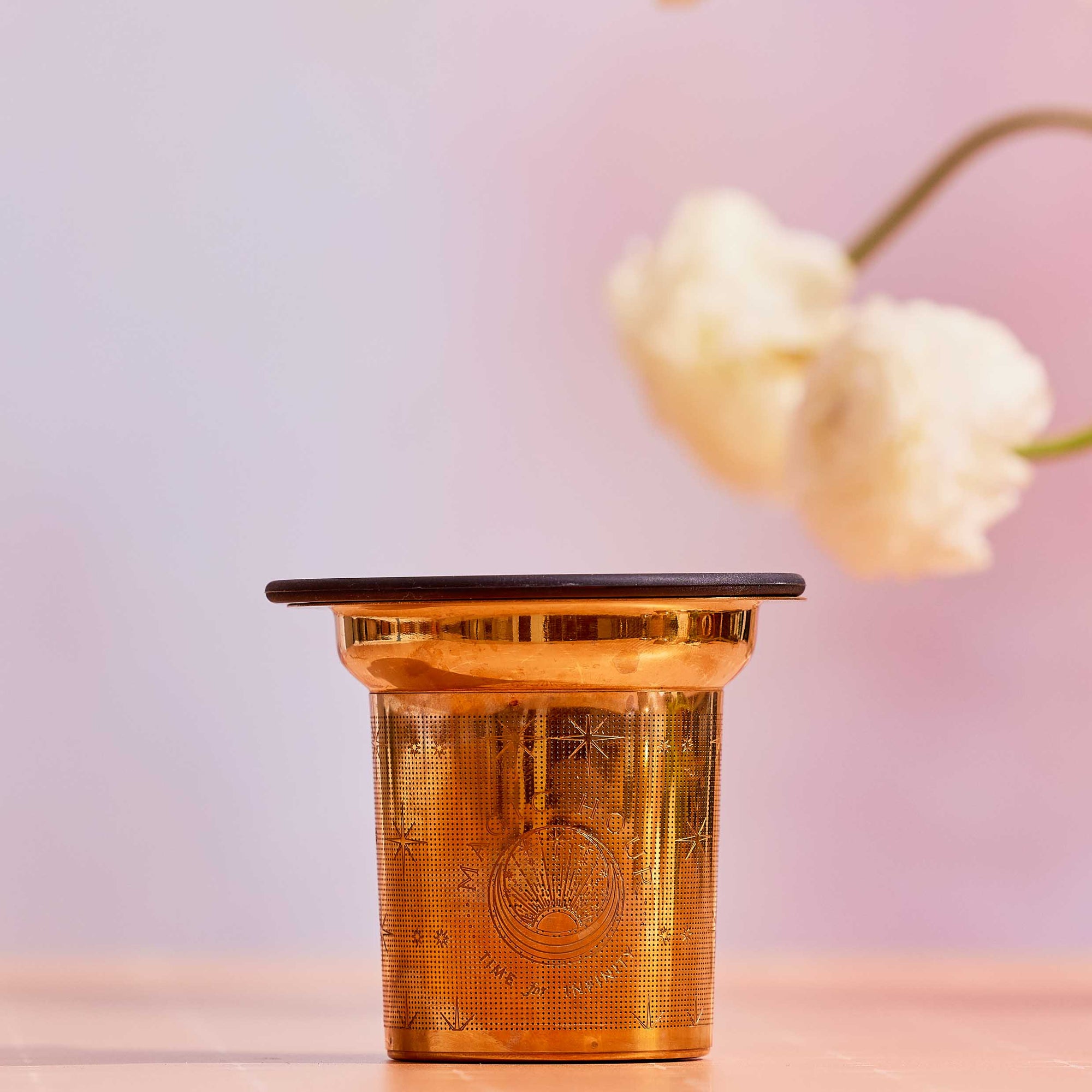 The Steep The Stars - Artisan Tea Strainer, featuring elegant engraved patterns, rests on a light pink surface with blurred white flowers and a soft pink backdrop, perfect for enjoying loose leaf tea.
