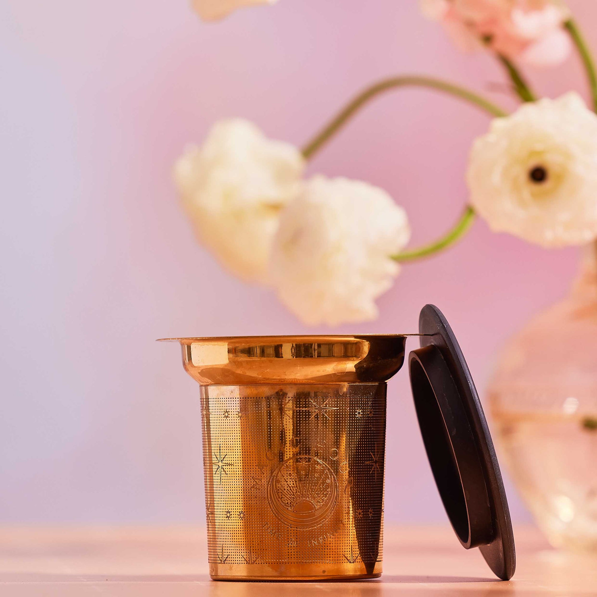 The Steep The Stars - Artisan Tea Strainer, a gold-toned infuser with a black lid, rests on a light surface beside a vase of white and pale pink flowers—ideal for brewing your favorite loose leaf tea.