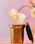 The Steep The Stars - Artisan Tea Strainer, a gold-toned infuser with a black lid, rests on a light surface beside a vase of white and pale pink flowers—ideal for brewing your favorite loose leaf tea.