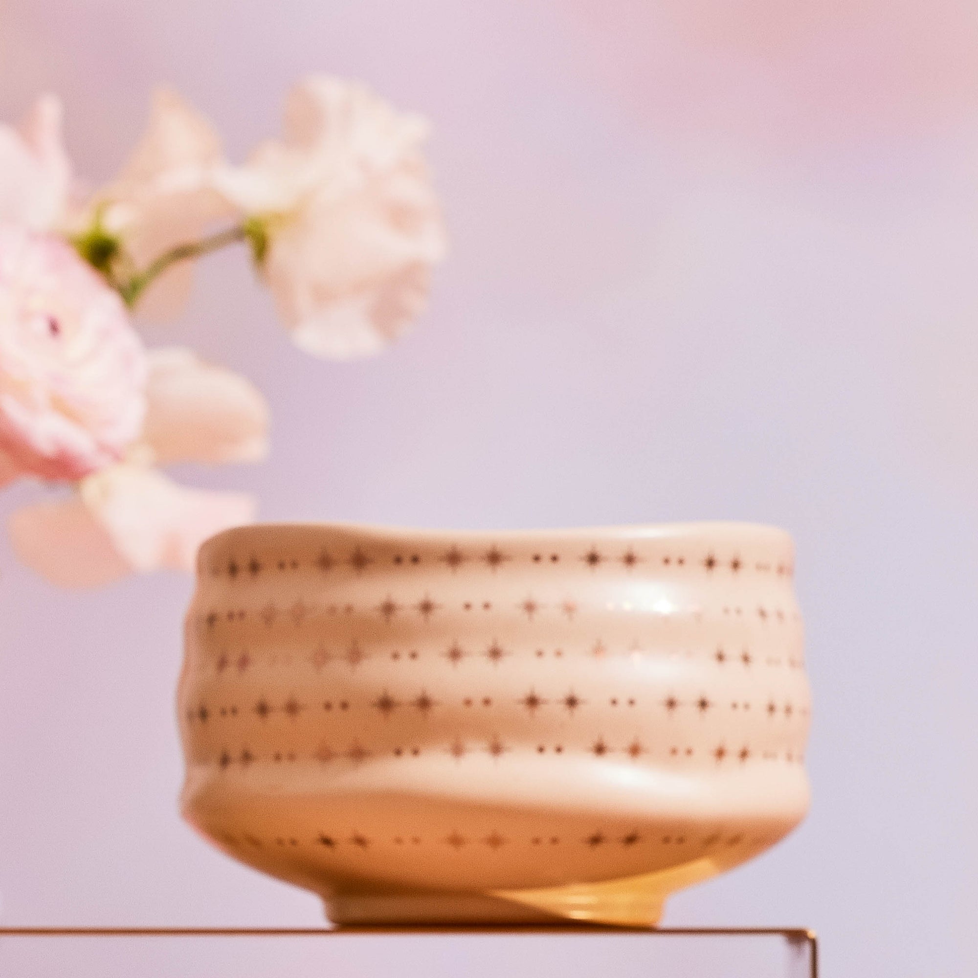 The Ceremonial Matcha Bowl, a handmade cream ceramic with star-shaped cutouts, rests on glass beside pink flowers against a pastel pink backdrop.