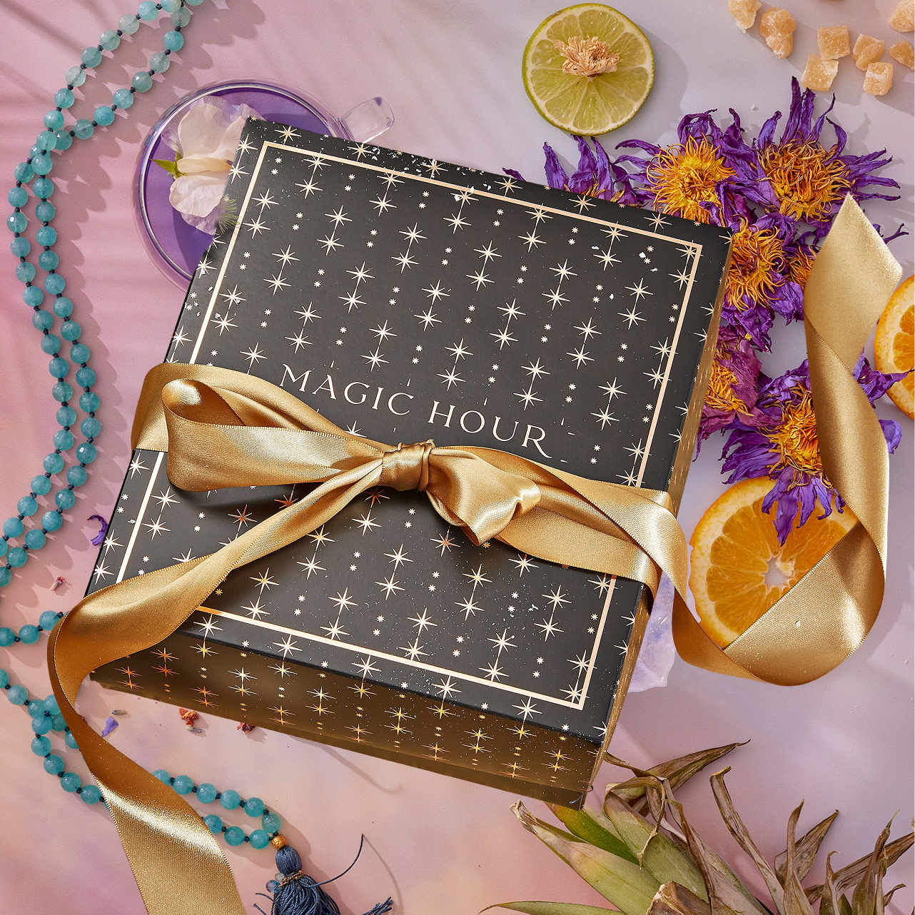 The Summer Solstice Mystery Box comes in a black gift box with gold stars and a gold ribbon labeled "Magic Hour," surrounded by dried flowers, turquoise beads, crystals, lime, orange slices, pineapple, and tea samplers.