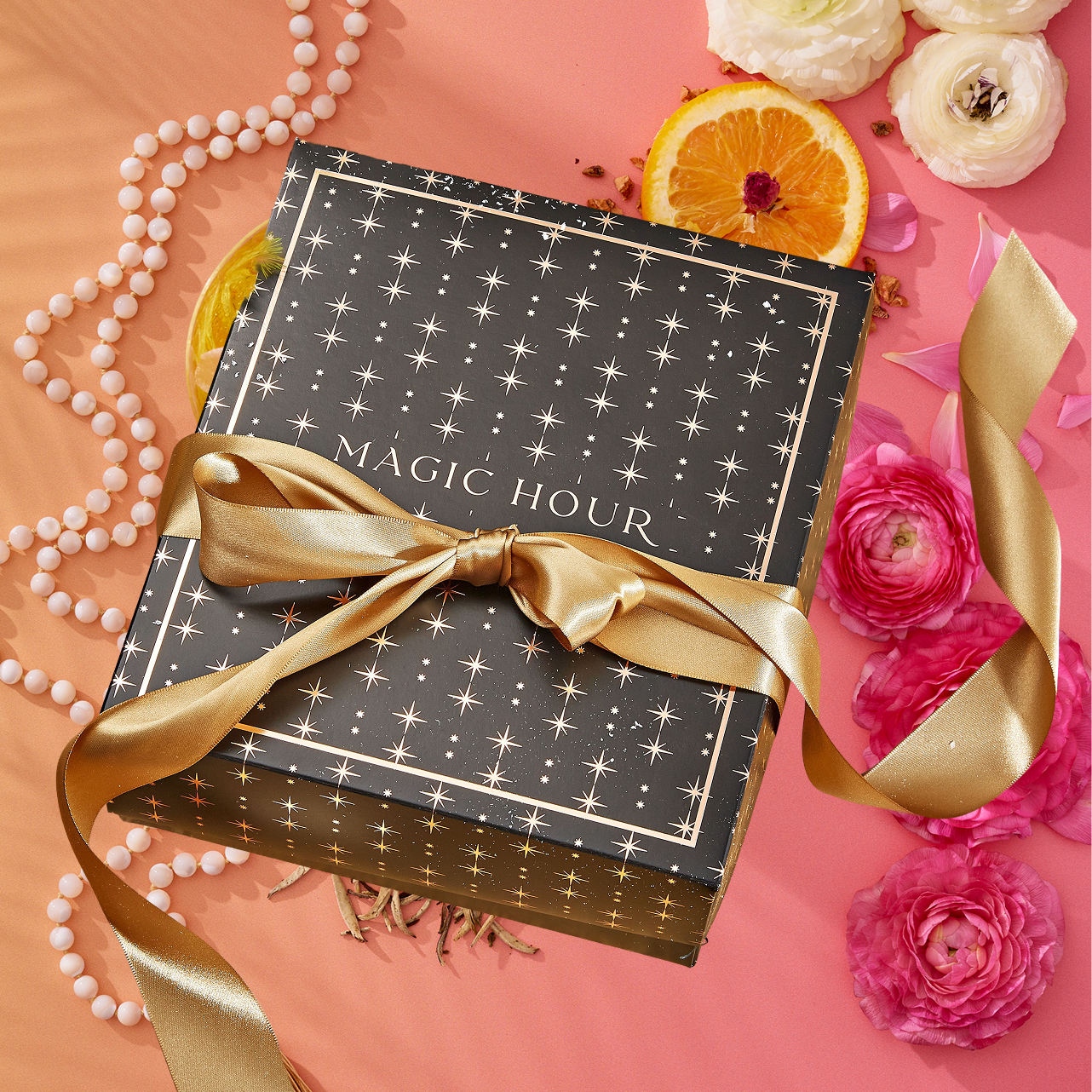 The Summer Solstice Mystery Box—a black gift box with gold stars and a "MAGIC HOUR" gold ribbon—is accented by pink flowers, an orange slice, and pearls. It includes exclusive, limited-edition tea samplers perfect for a magical summer surprise.