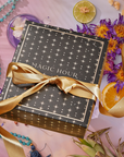 The Summer Solstice Mystery Box comes in a black gift box with gold stars and a gold ribbon labeled "Magic Hour," surrounded by dried flowers, turquoise beads, crystals, lime, orange slices, pineapple, and tea samplers.