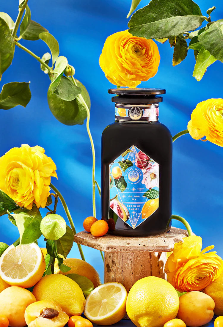 A dark bottle with a colorful, diamond-shaped label is placed on a cork pedestal surrounded by vibrant yellow flowers, green leaves, and various citrus fruits including lemons and kumquats. The background features a bright blue fabric backdrop.