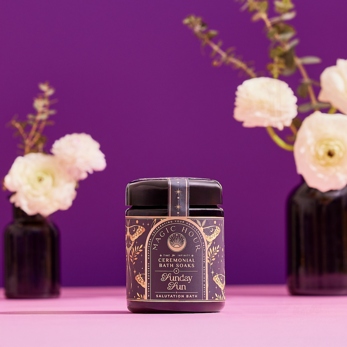 A Violet Glass Jar of Ceremonial Bath Salt Soaks is showcased on a pink surface, with dark vases of white flowers and greenery against a vibrant purple background, creating a celestial vibe.