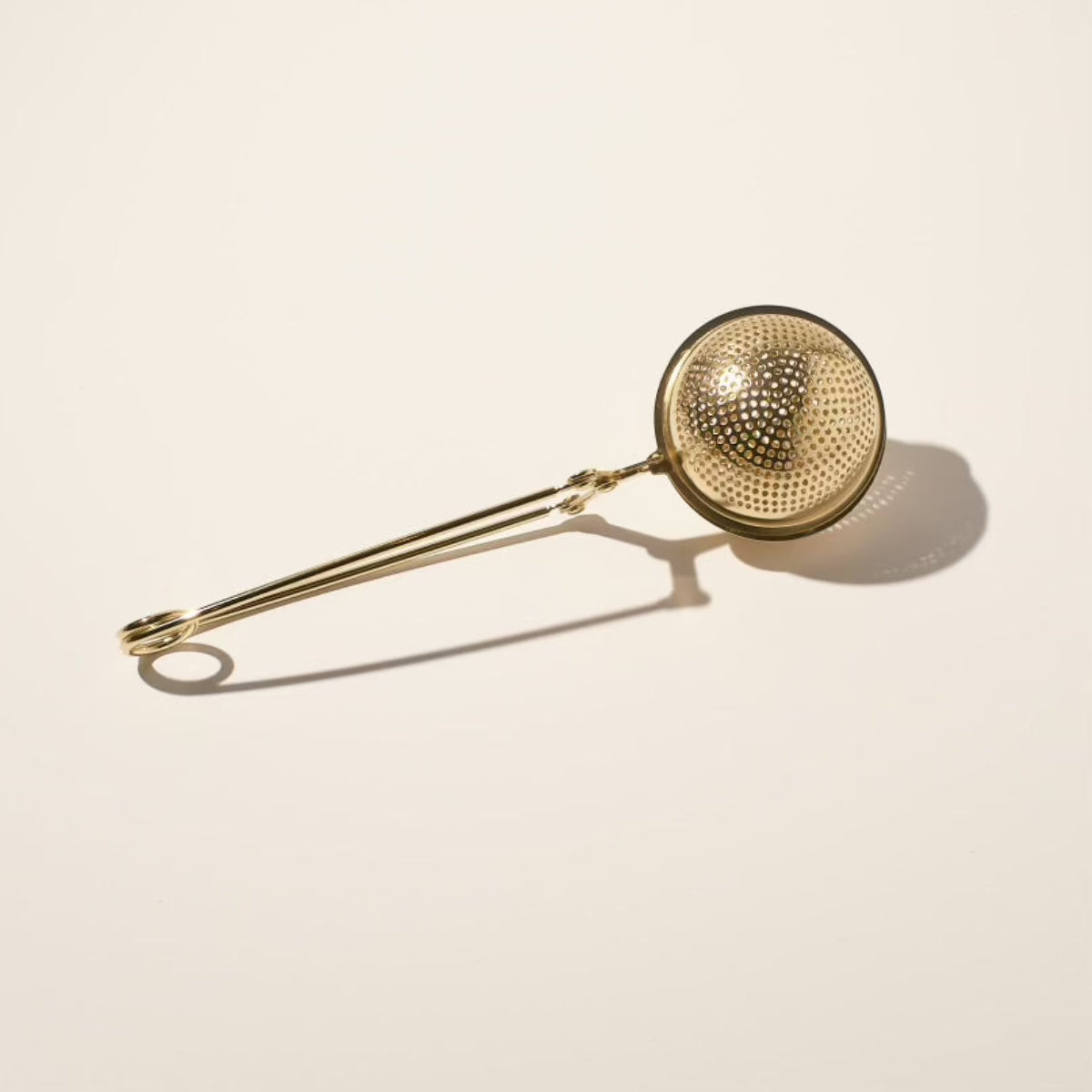 A Magic Hour Starter Kit features a gold-colored metal tea infuser with a spherical mesh strainer and a long handle, beautifully showcased on a light beige background. Ideal for artisan-crafted strainers, the handle includes a small loop at the end. The shadow of the infuser is visible, suggesting light coming from the left.