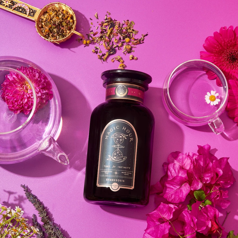 A beautiful display featuring a jar of Symbeeosis: Beautifying Immunitea for the Queen Bee on a vibrant pink background. Surrounding the jar are a transparent teapot, two transparent cups, a spoon with loose leaf tea, and assorted colorful flowers. This serene setup invites you to start your wellness journey with health-boosting ingredients.