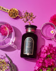 A beautiful display featuring a jar of Symbeeosis: Beautifying Immunitea for the Queen Bee on a vibrant pink background. Surrounding the jar are a transparent teapot, two transparent cups, a spoon with loose leaf tea, and assorted colorful flowers. This serene setup invites you to start your wellness journey with health-boosting ingredients.